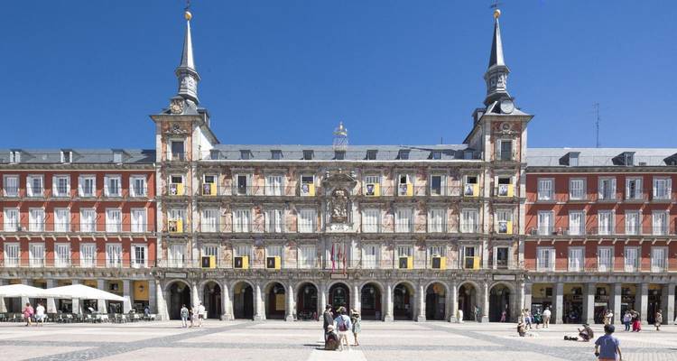Symmetrical facade of Madrid's Plaza Mayor bathed in sunlight with visitors strolling the square.