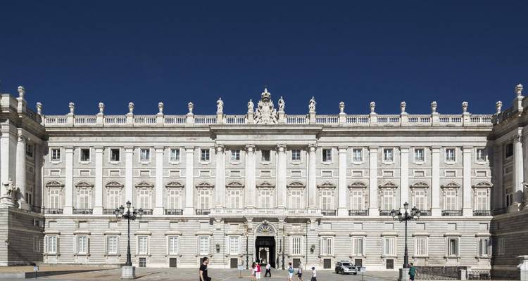 Frontal view of the grand Royal Palace with tiny visitors dotting the expansive forecourt under a deep blue sky