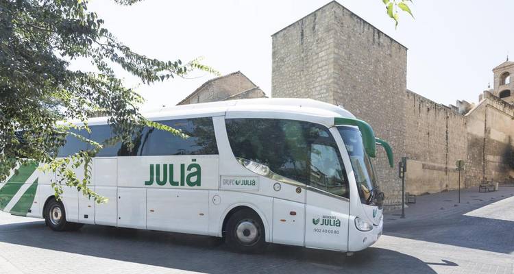 Un moderno autobús turístico blanco de marca 'Juliá' estacionado junto a un antiguo edificio de piedra en un día soleado.