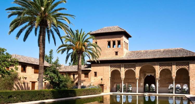 El Palacio del Partal de la Alhambra reflejado en una piscina tranquila, enmarcado por palmeras bajo cielos azules despejados.