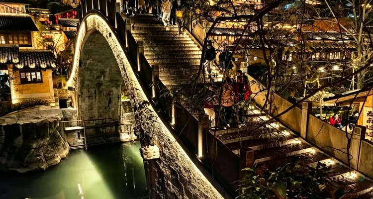 Puente de arco de piedra y escaleras iluminadas por lámparas cálidas con visitantes del atardecer cruzando un canal estrecho.