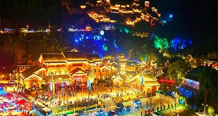 Vista nocturna vibrante de una plaza animada y un pueblo de montaña iluminados con luces de múltiples colores.