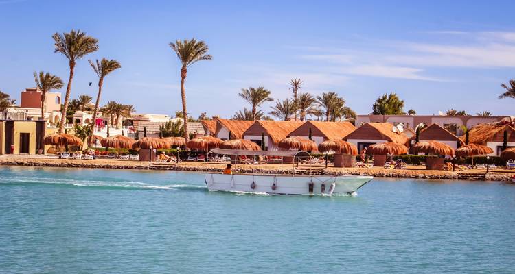 Station balnéaire avec lagon, palmiers et cabanas aux toits de chaume tandis qu'un bateau navigue sur une eau bleue et calme
