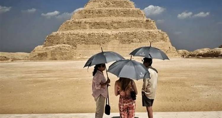 Drei Besucher unter Regenschirmen stehen an einem sonnigen Tag vor der ikonischen Stufenpyramide von Saqqara.
