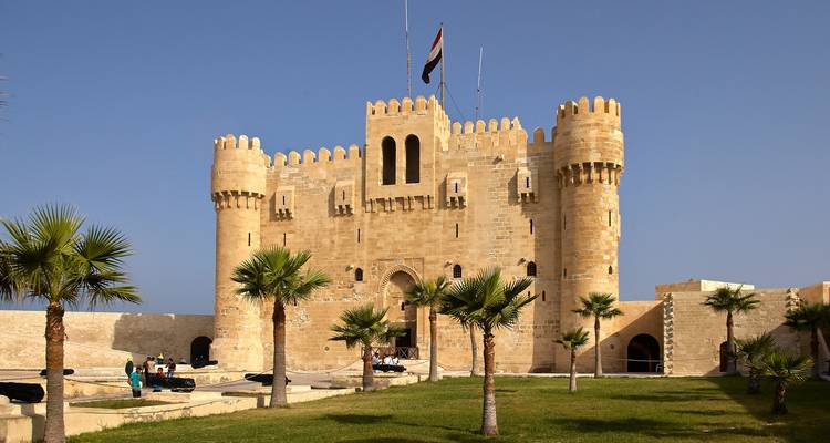 Sunlit view of the medieval Qaitbay Citadel overlooking the Mediterranean with tourists nearby.
