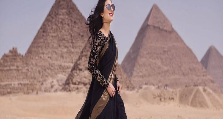 Fashionable woman in black sari smiling while walking near Giza pyramids.