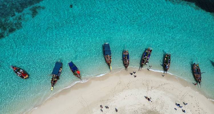 Aerial view of traditional wooden boats lined up on brilliant turquoise water beside a white sandy beach.