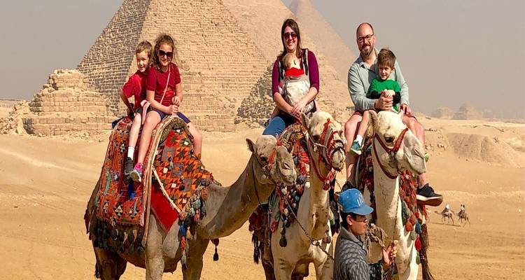Una familia pasea en dos camellos coloridos y decorados frente a las pirámides en un día nublado.
