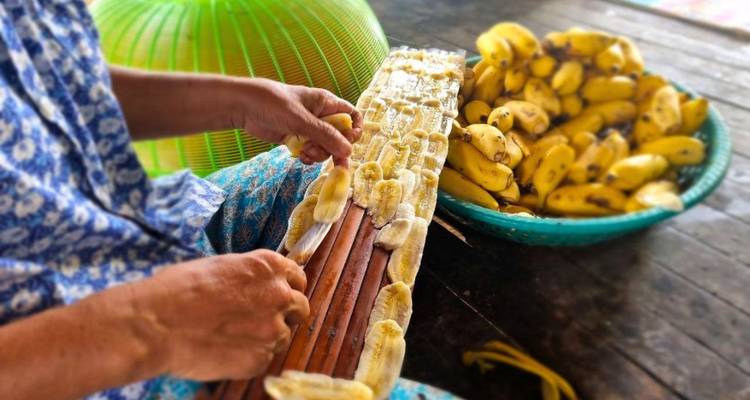 Primer plano de una mujer colocando plátanos rebanados en una tabla de madera al lado de una canasta de plátanos pequeños.