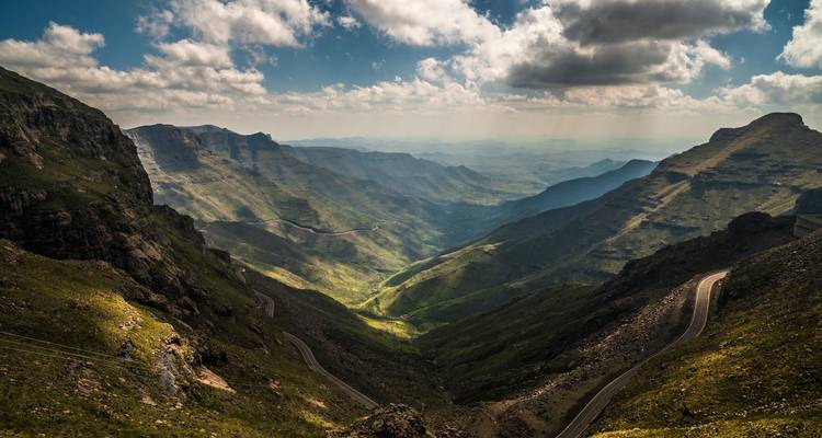 Col de montagne spectaculaire avec route sinueuse descendant à travers des vallées vertes abruptes sous des nuages épars.
