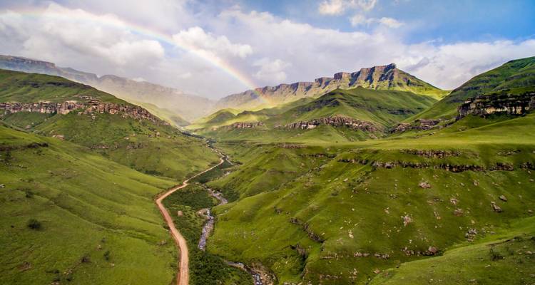 Des montagnes verdoyantes luxuriantes et une route de vallée mises en évidence par un arc-en-ciel éclatant s'arquant à travers un ciel partiellement nuageux.