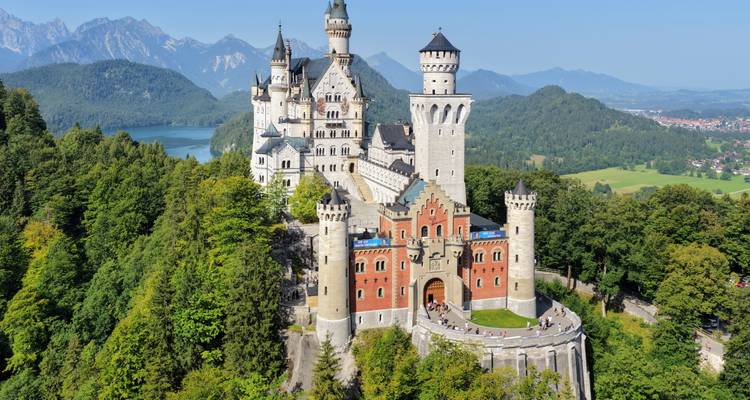 El icónico Castillo de Neuschwanstein encaramado sobre colinas boscosas con un lago alpino y montañas de fondo.