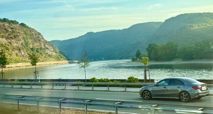 Un coche circulando por una carretera junto al río con colinas verdes y agua tranquila en un día soleado.