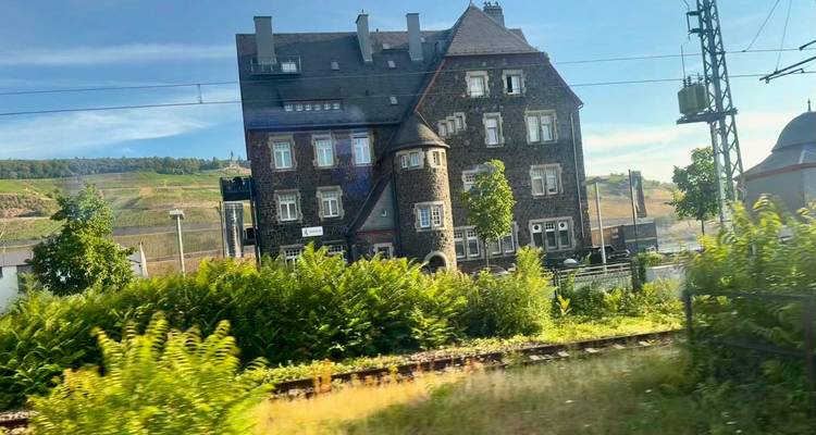 Vista borrosa desde la ventana del tren de un edificio de piedra y vías de ferrocarril rodeadas de vegetación bajo un cielo despejado.