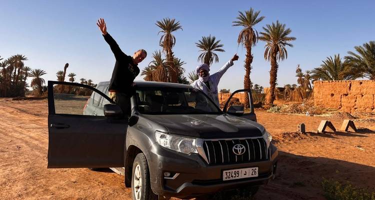 Deux voyageurs célébrant à côté d'un 4x4 entouré de palmiers et de murs en adobe dans le désert