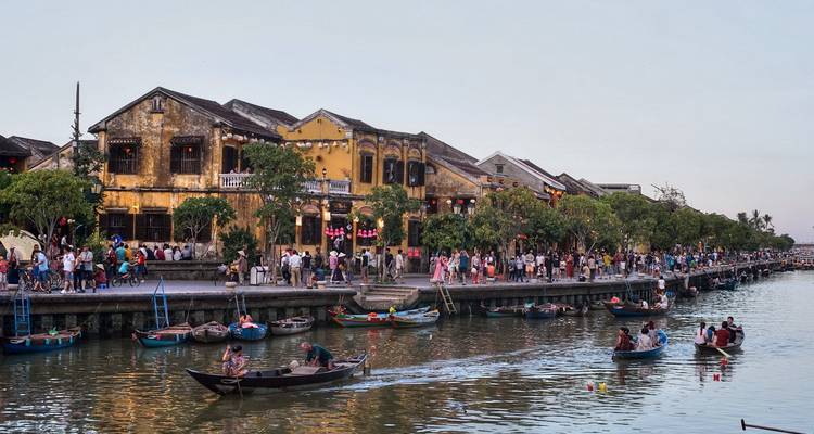 Menschenmengen spazieren entlang der Ufergegend von Hoi An, wo Boote neben verwitterten gelben Geschäftshäusern im Licht des späten Nachmittags schwimmen.