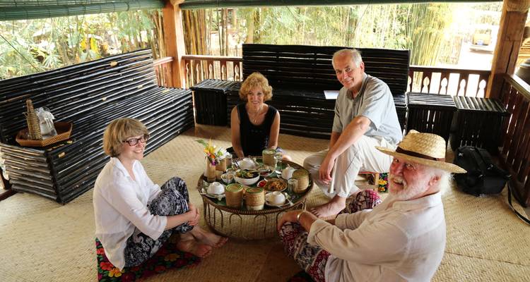 Cuatro visitantes se sientan con las piernas cruzadas alrededor de una mesa baja compartiendo un almuerzo laosiano en un salón de bambú.
