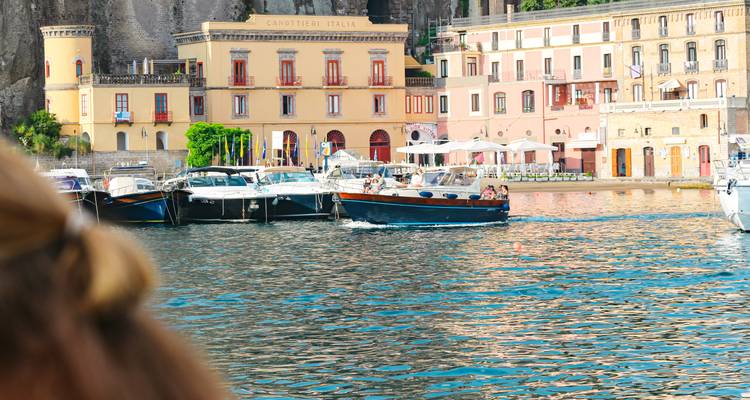 Havenschene in Sorrento met boten aangemeerd onder pastelkleurige gebouwen aan de waterkant; een wazig figuur vormt de voorgrond.
