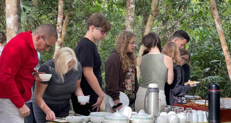 Travelers serving themselves breakfast from outdoor buffet in a forest clearing