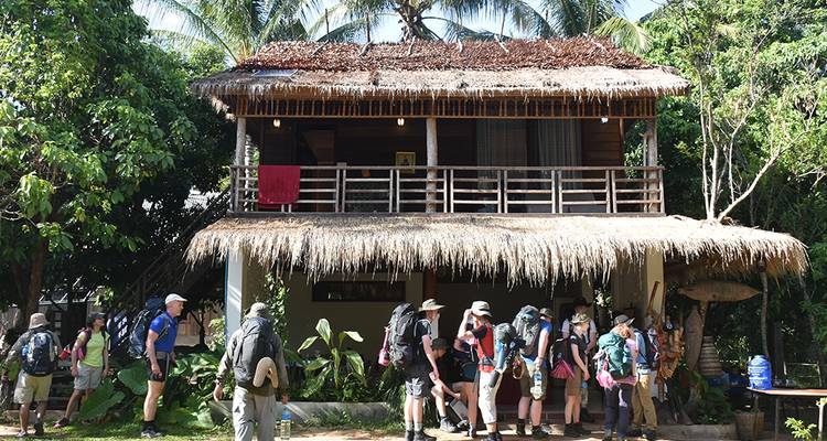 Backpack-laden hikers gathering outside a rustic wooden lodge with thatched roof