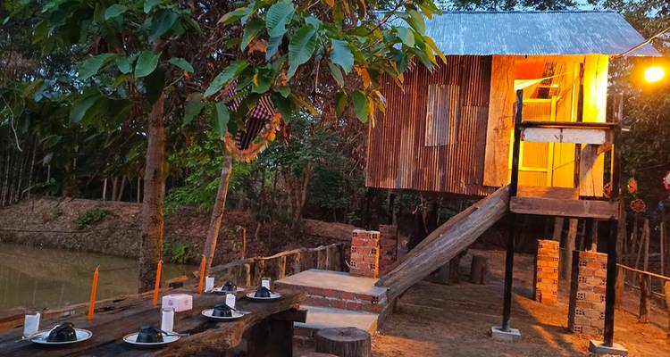 Stilted wooden cabin lit warmly at dusk beside an outdoor dining bench