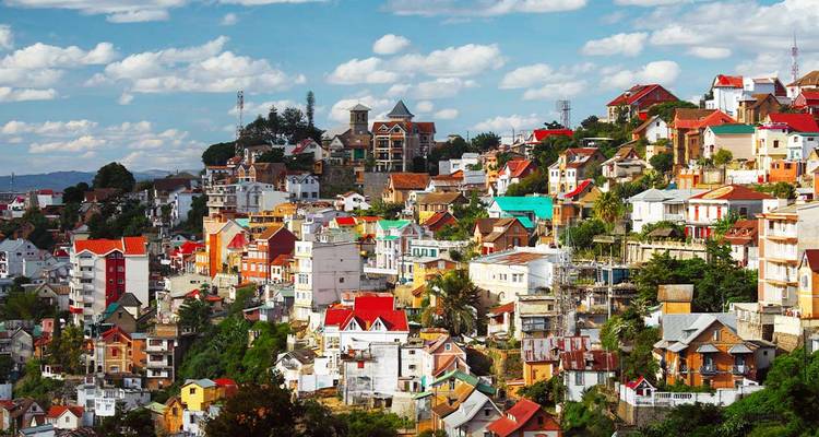 Des maisons colorées s'empilent sur les collines ondulantes d'Antananarivo sous un ciel lumineux parsemé de nuages.