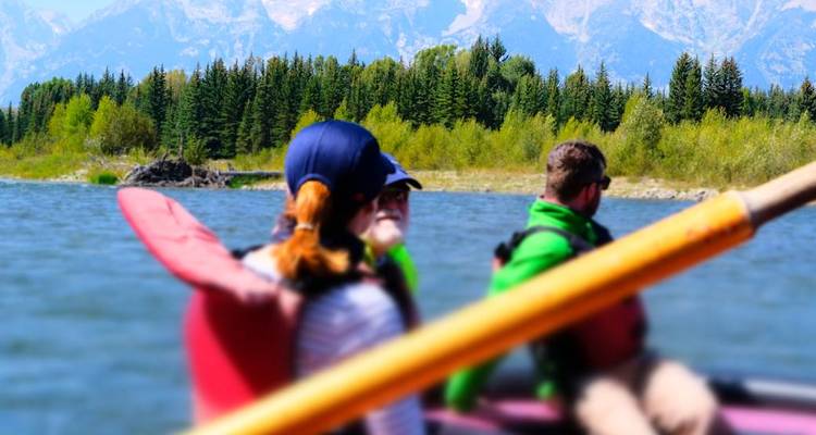 Groupe pratiquant le rafting sur une rivière calme avec une pagaie floue au premier plan et une forêt de pins et des montagnes à l'arrière-plan.