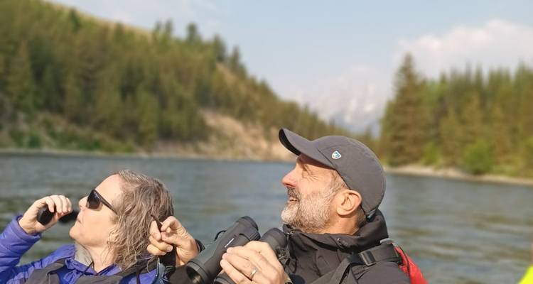 Deux voyageurs assis dans un radeau gonflable observent la forêt riveraine à travers des jumelles par une journée dégagée.
