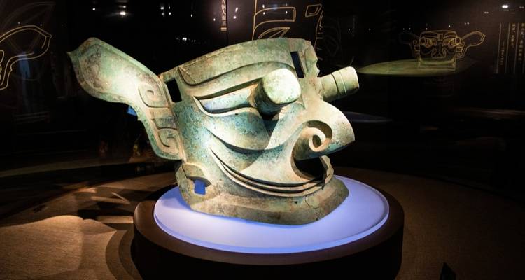 Een oud bronzen Sanxingdui maskerartefact is dramatisch verlicht in een donkere museumvitrine.