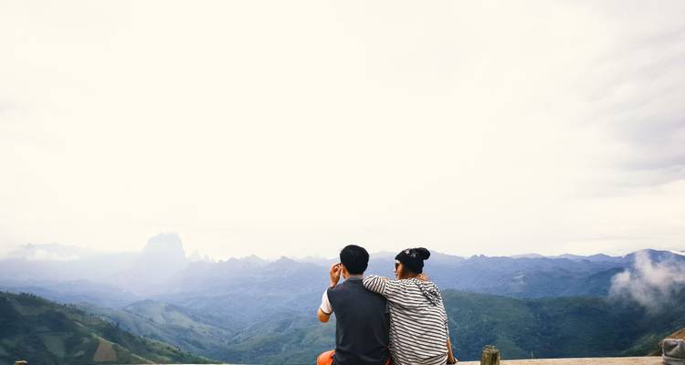 Ein Paar sitzt auf einer Holzplattform mit Blick auf neblige Bergketten und Täler, die sich bis zum Horizont erstrecken.