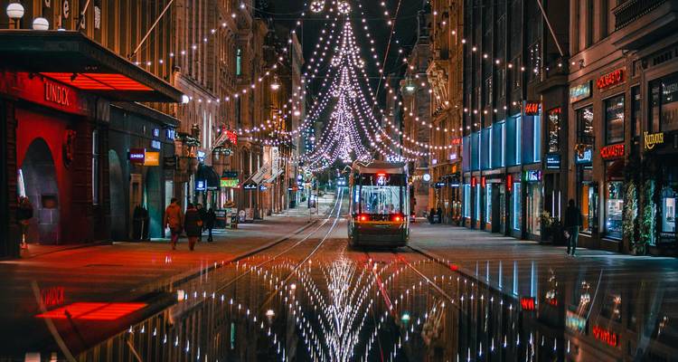 Nighttime city street in Helsinki with a tram approaching under strings of festive lights, reflections shimmering on wet pavement.