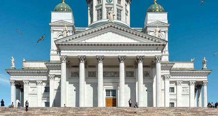 Frontal view of the white-columned Helsinki Cathedral framed by a bright blue sky, a few visitors on the wide stone steps.