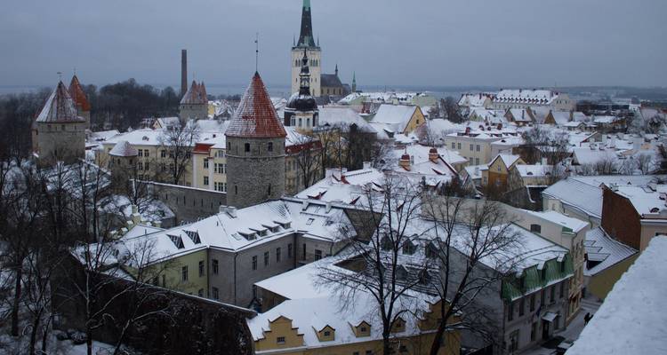 Snow-covered medieval towers and church spires of Tallinn under a grey winter sky seen from a high vantage point.