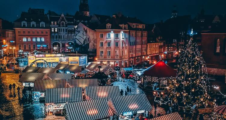 Bird’s-eye nighttime view of a lively Christmas market with striped vendor stalls, twinkling lights and crowds milling about.
