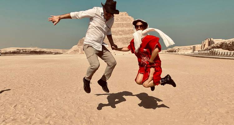 Un homme et une femme en saut suspendu dans les airs devant la Pyramide à Degrés de Saqqarah lors d'une matinée désertique ensoleillée.