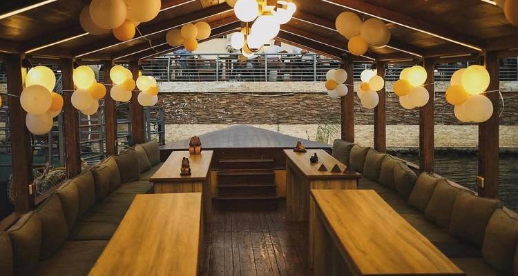 Een gezellig houten bootinterieur met gestoffeerde zitjes en warm licht dat feestelijk is versierd met gele ballonnentrossen terwijl het afgemeerd ligt in het water.