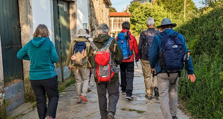 Groep pelgrims met rugzakken wandelen langs een zonnig landweggetje tussen stenen gebouwen.
