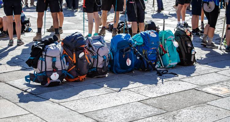 Rij met kleurrijke trekrugzakken liggen op de grond terwijl pelgrims op een zonnig plein staan.