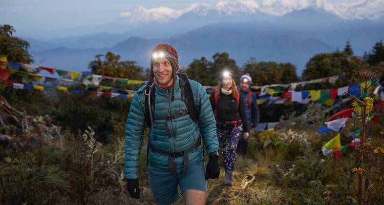 Trekking group with headlamps hiking at dawn amid Himalayan prayer flags and snowy peaks.