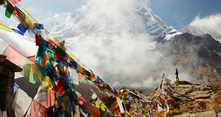 Prayer flags flutter toward snow-clad peaks shrouded in swirling clouds with a lone trekker.