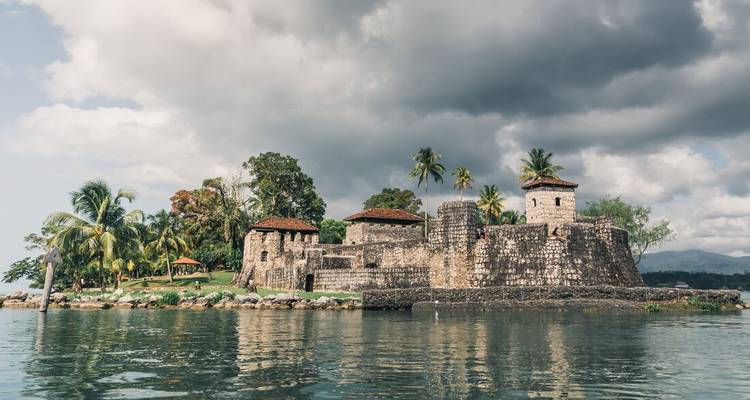 Un fort colonial en pierre se dresse au bord des eaux calmes d'une rivière bordée de palmiers sous des nuages dramatiques.