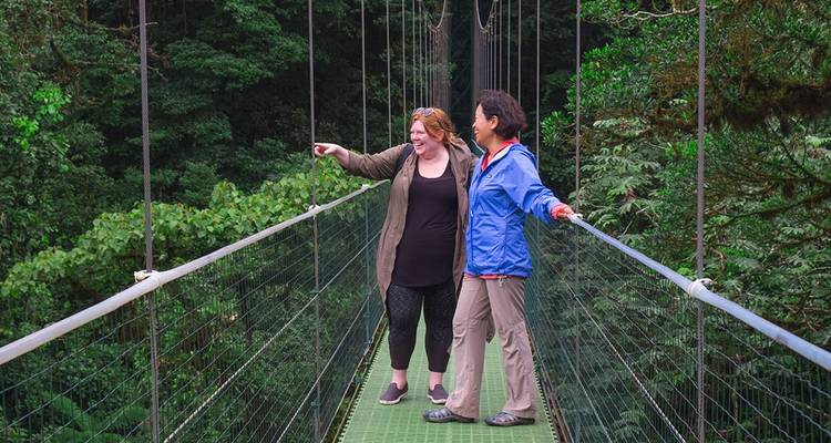 Two young women stand on a suspended canopy bridge overlooking dense rainforest, one pointing ahead.