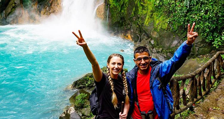 Smiling man and woman flash peace signs beside a turquoise waterfall set in lush jungle.