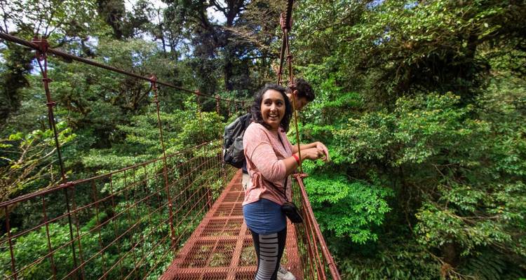 Un voyageur sourit en traversant un pont suspendu en métal rouge situé haut dans une forêt nuageuse.