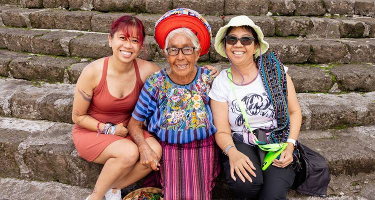Deux voyageurs sont assis avec un aîné maya traditionnellement vêtu sur des marches de pierre anciennes, tous souriants.