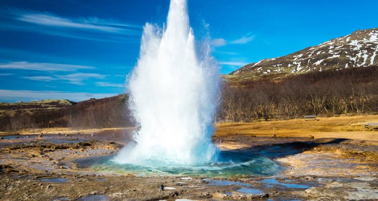 Strokkur geyser erupts high into a blue sky amid Icelandic volcanic landscape.