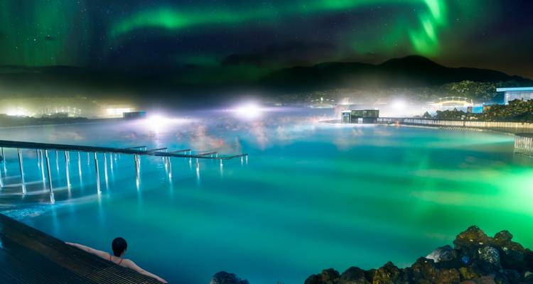Steaming turquoise Blue Lagoon under northern lights with a lone bather enjoying the geothermal pool.