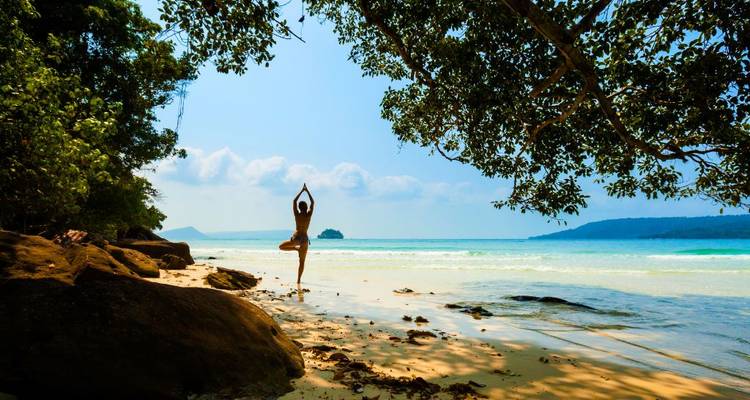 Silueta de una mujer practicando yoga en una tranquila playa tropical con agua turquesa y árboles colgantes.