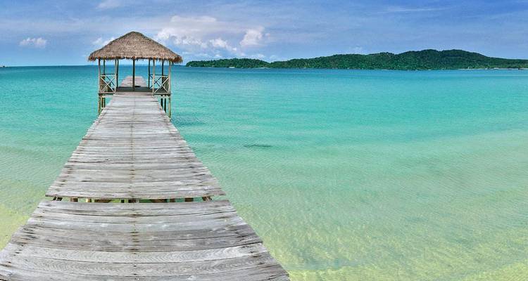 Largo muelle de madera con una cabaña de paja extendiéndose sobre un mar turquesa cristalino hacia una isla verde distante.
