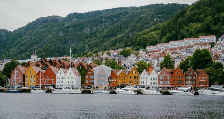 Fila de casas coloridas de Bryggen reflejadas en el agua tranquila del puerto con una ladera boscosa de fondo.
